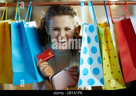 heureuse femme d'âge moyen élégante en sweat blanc et jupe se démontant de sacs de shopping colorés accrochés sur rail de vêtements en cuivre avec carte de crédit rouge Banque D'Images