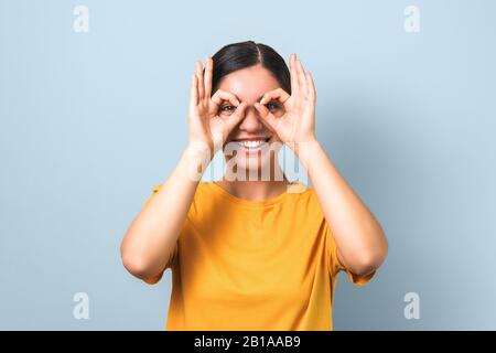 belle brunette en t-shirt jaune faisant des verres de ses doigts et visage souriant drôle sur fond bleu Banque D'Images