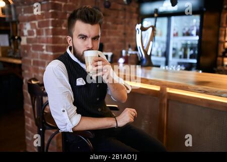 jeune homme sérieux dans des vêtements élégants buvant une nouvelle sorte de café, arôme qui n'est pas résisté par un barmen Banque D'Images