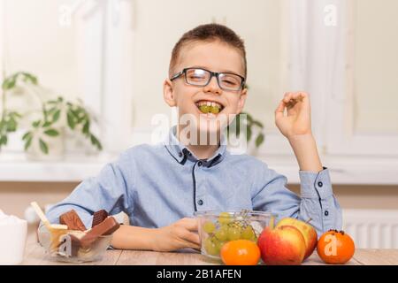 Un garçon souriant se trouve à la table et mange des fruits. Il a des fruits de raisin dans ses dents. Banque D'Images