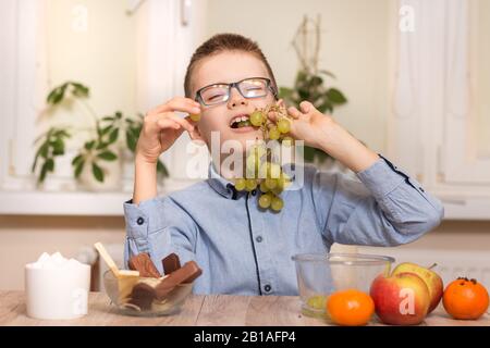 Un garçon souriant se trouve à la table et mange des fruits. Il a des fruits de raisin dans ses dents. Banque D'Images