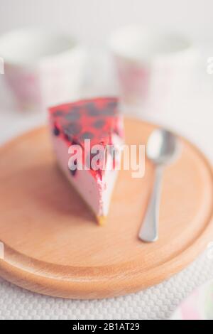 Un morceau de cheesecake de curricuille et une cuillère sur une planche ronde en bois. Deux tasses de thé sur une table blanche sur fond flou. Gâteau au fromage rose avec noir Banque D'Images