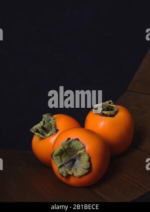 Хурма. Persimmons sur fond sombre. Persimmons sur une table en bois. Trois persimmons mûrs sur un fond sombre. Banque D'Images