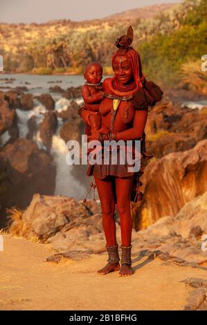 Himba femme sport son cou plein d'ornements traditionnels et un petit bébé sur ses genoux, Epupa Falls en arrière-plan, région de Kunene, Namibie Banque D'Images