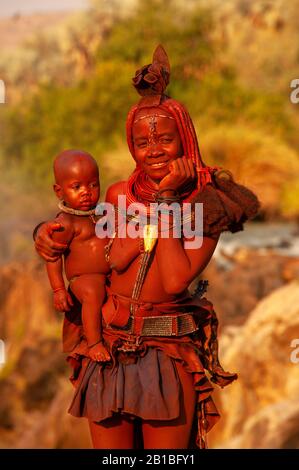 Himba femme sport son cou plein d'ornements traditionnels et un petit bébé sur ses genoux, Epupa Falls, région de Kunene, Namibie Banque D'Images