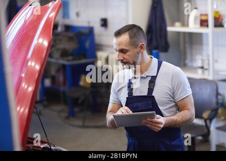 Taille vers le haut portrait du mécanicien de voiture barbu utilisant la tablette numérique pendant l'inspection du véhicule dans le garage, espace de copie Banque D'Images