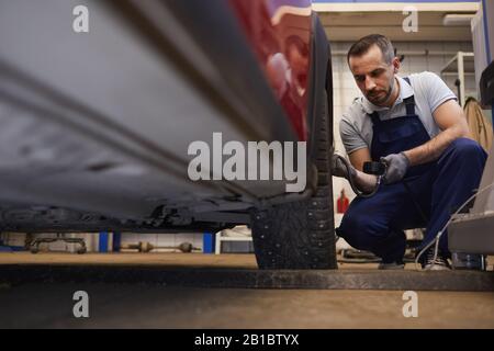 Portrait à bas angle du mécanicien de voiture barbu vérifiant la pression dans les pneus pendant l'inspection du véhicule dans l'atelier de garage, espace de copie Banque D'Images