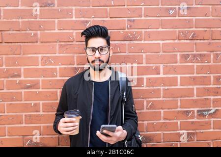 Beau homme indien utiliser le téléphone mobile en profitant du café contre le mur de briques Banque D'Images