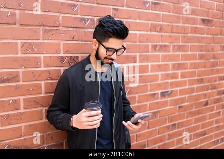 Beau homme indien utiliser le téléphone mobile en profitant du café contre le mur de briques Banque D'Images