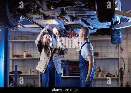 Portrait de deux mécaniciens matures se tenant sous la voiture au levage tout en inspectant le véhicule dans un atelier de réparation automatique, espace de copie Banque D'Images