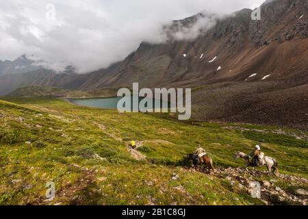 Plusieurs personnes à cheval et marchez le long d'une vallée verte dans les montagnes jusqu'au lac. Chevaux chargés de balles. Nuages au-dessus des hautes montagnes. Banque D'Images