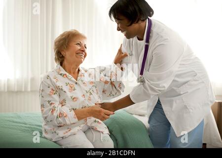 Smiling senior woman holding la main d'une femme médecin alors qu'elle se trouve sur le bord d'un lit à l'intérieur d'une chambre. Banque D'Images