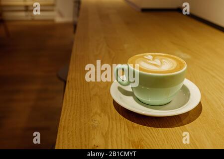 Regardez l'art du Latte avec le motif coeur sur le café cappuccino sur une tasse en céramique vert pâle dans le café intérieur du comptoir en bois. Banque D'Images