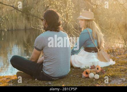 Jeune couple assis sur l'herbe devant la rivière au coucher du soleil. Relations humaines, émotions positives Banque D'Images