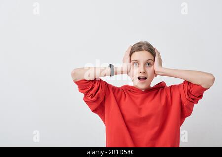 Portrait d'une adolescente choquée dans un sweat à capuche rouge en gardant la bouche ouverte et en tenant la tête dans les mains contre un fond blanc Banque D'Images