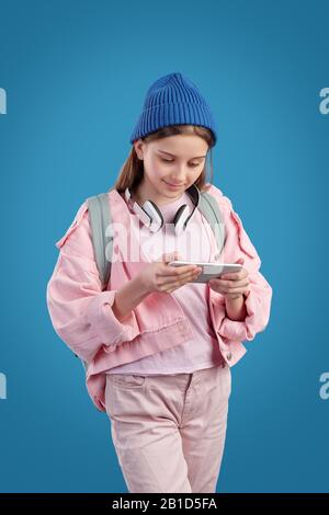 Curieuse fille adolescente dans une veste rose portant un casque sans fil sur le cou regardant la vidéo au téléphone Banque D'Images