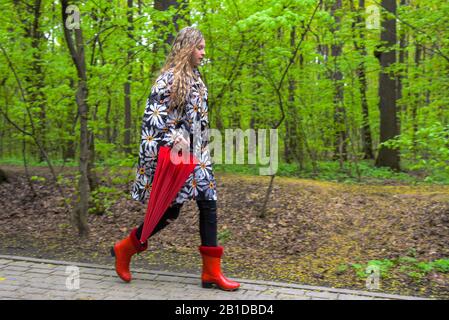 Une adolescente avec de longs cheveux blonds portant des bottes en caoutchouc rouge et un parapluie rouge marche le long d'une allée déserte dans le parc du printemps. Banque D'Images