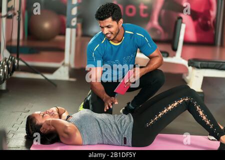 L'entraîneur aide la jeune femme à faire des exercices abdominaux sur le fond des balles de fitness. Belle femme faisant l'abs sur un tapis et un homme l'aide dans Banque D'Images