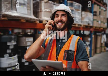 Employé de l'entrepôt utilisant une tablette numérique pendant une conversation sur un téléphone mobile dans un entrepôt portant un casque blanc et un gilet de sécurité Banque D'Images