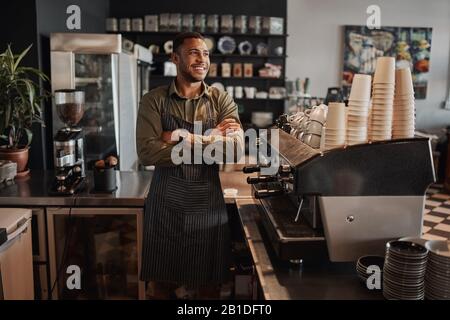 Petit propriétaire afro-américain attentif se tenant derrière le comptoir portant un tablier avec des bras croisés regardant loin Banque D'Images