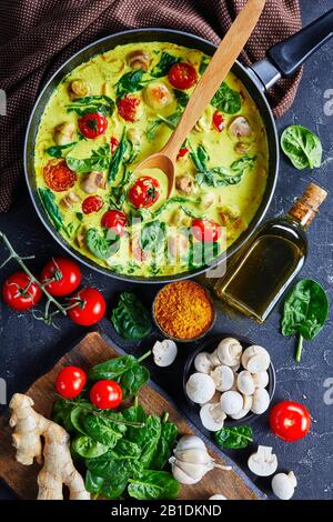 Curry indien sur une poêle de lait de coco aux champignons et légumes : épinards et tomates cerises sur fond de béton foncé avec ingrédients, t Banque D'Images