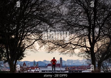 Munich, Allemagne. 25 février 2020. Un jogger se dresse sur la colline de Luitpold, dans le quartier de Schwabing, et regarde sur la toile de fond de la capitale bavaroise, dont le centre est marqué par les tours de 99 mètres de haut de la Frauenkirche (centre droit). Derrière les gratte-ciel de la ville, vous pourrez admirer le panorama alpin légèrement enneigé. La distance entre le monument et les Alpes est d'environ 100 kilomètres et ne peut être appréciée que par temps foehn distinct. Crédit: Peter Knelove/Dpa/Alay Live News Banque D'Images