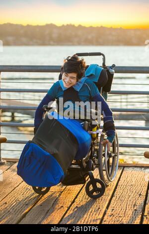 Petit garçon biracial handicapés assis en fauteuil roulant sur la jetée en bois au bord du lac au coucher du soleil Banque D'Images