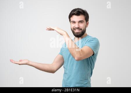 Jeune homme hispanique avec barbe tenant deux mains devant lui et montre la taille sur fond blanc isolé en studio. Banque D'Images