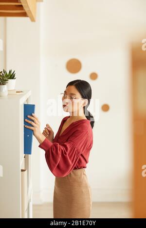 Vue latérale portrait d'une élégante femme d'affaires asiatique mettant la reliure à la bibliothèque contre le mur blanc au bureau, espace de copie Banque D'Images