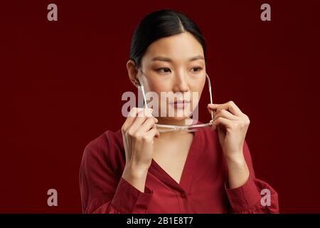 Tête et épaules portrait d'une élégante femme asiatique mettant sur des lunettes tout en se tenant sur fond de marron en studio, espace de copie Banque D'Images