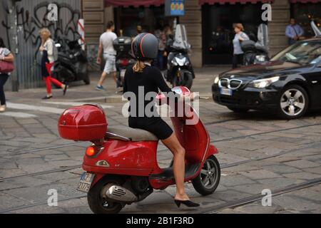 Une femme qui monte un scooter rouge à moteur dans la circulation portant une robe noire et des mules en daim noir, Porsche Design noir et casque rouge sur Via Giuseppe Mazzini Banque D'Images