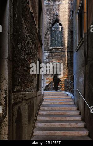 Venise, une calle étroite, marches en marbre blanc sur un pont avec balustrade ornée de dentelle de fer, fenêtre gothique à volets dans un ancien mur de briques et une femme Banque D'Images