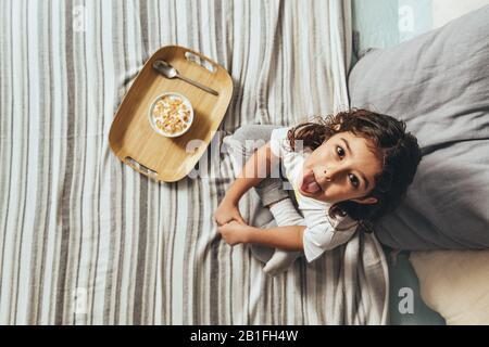 vue de dessus d'une petite fille taqueuse qui s'enfile dans sa langue avec un petit déjeuner le dimanche dans le lit de ses parents. Enfant profitant d'un bol de céréales à la maison, concept Banque D'Images