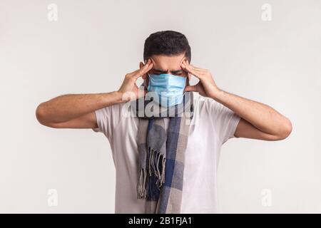 Homme malsain dans le masque chirurgical et le foulard souffrent de maux de tête, frottant des temples douloureux, ayant des symptômes de grippe, infection contagieuse de coronavirus 2019-NC Banque D'Images