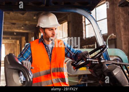 Workman porte un casque de protection, un gilet et des gants. Travailleur debout près du chariot élévateur à fourche dans l'entrepôt. Banque D'Images