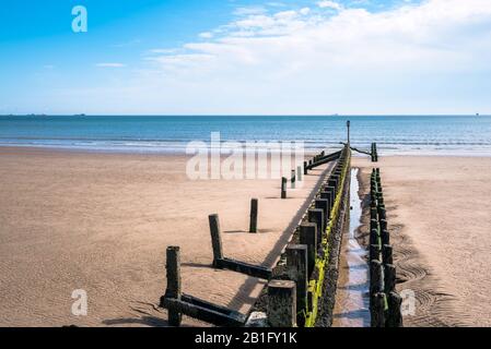 Groyne sur une plage de sable sur une matinée de printemps partiellement nuageux. Certains navires ainsi que des éoliennes offshore sont visibles à l'horizon. Banque D'Images