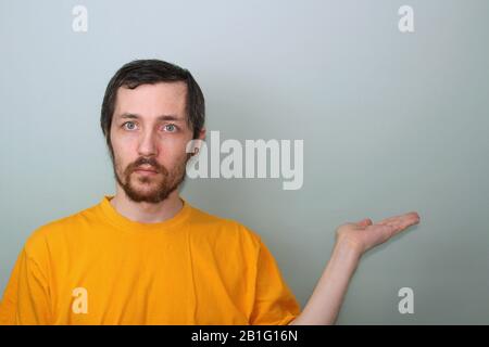 Un homme d'âge moyen avec une moustache et une barbe brunette dans un T-shirt jaune sur fond gris Tient la main de côté à paume vers le haut avec un espace libre au-dessus de la paume. Banque D'Images