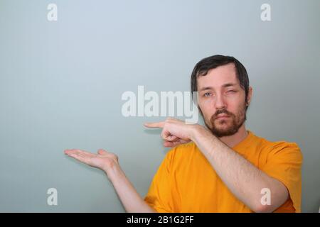 Un homme d'âge moyen avec une moustache et une barbe brunette dans un T-shirt jaune sur fond gris Tient la main sur le côté, la paume vers le haut avec l'espace libre au-dessus de la paume, pointe un doigt sur l'espace au-dessus de la paume et se calque les yeux. Banque D'Images