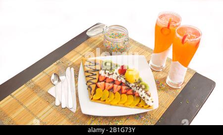 Vue de dessus de la crêpe avec des tranches de fraise et de pêche garnies de chocolat, crème et crème glacée aux fruits sur une plaque blanche avec deux tasses de verre avec jus Banque D'Images