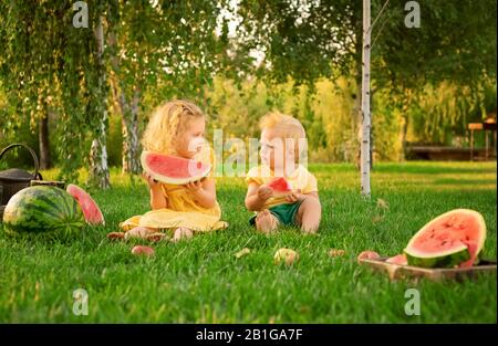 Joyeux enfant souriant mangeant du pastèque à l'extérieur lors d'un pique-nique en été au coucher du soleil. Bébé et fille blonde blanche, sœur et frère mangeant du pastèque Banque D'Images