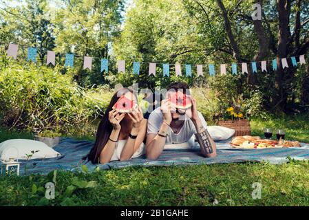 Jeune couple hétérosexuel posé sur le sol sur une couverture ayant pique-nique et regardant par un morceau de pastèque sélective foyer Banque D'Images