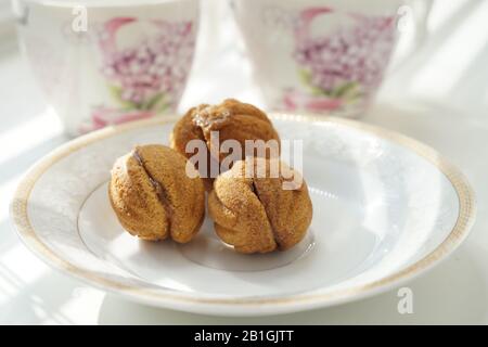 Trois petits cookies sous forme de noix sur une soucoupe blanche. Arrière-plan deux tasses de thé sur une table ensoleillée. Banque D'Images