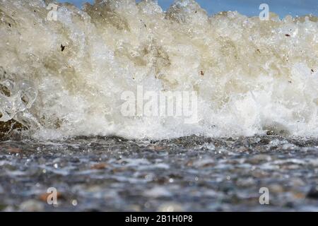 Gros plan sur une petite vague de rupture sur une plage. Certains débris sont visibles. Vue en angle bas depuis l'avant avec le sol en regardant directement la vague. Banque D'Images