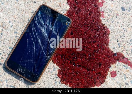 Un téléphone portable cassé et écrasé par une flaque de sang sur un trottoir. L'écran du téléphone portable est cassé et brisé. Le sang est frais. Banque D'Images