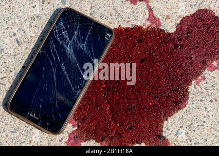Un téléphone portable cassé et écrasé par une flaque de sang sur un trottoir. L'écran du téléphone portable est cassé et brisé. Le sang est frais. Banque D'Images