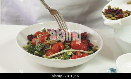 Préparation de la salade de légumes. Femme met des graines de citrouille et des canneberges séchées dans un bol avec des feuilles de salade et des tomates, gros plan sur le dos blanc Banque D'Images