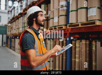 Jeune employé en entrepôt dans un casque et une veste de sécurité utilisant une tablette numérique tout en prenant commande et en confirmant la disponibilité des stocks à l'aide d'une tablette numérique Banque D'Images