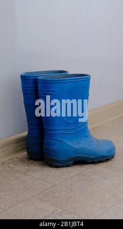 Une paire de bottes en caoutchouc bleues pour enfants se tenant le long d'un mur blanc sur le linoléum Banque D'Images