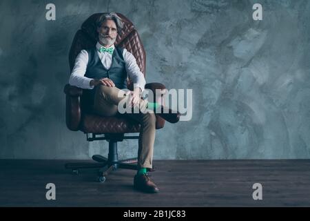 Photo pleine longueur de l'élégant masculine ancien propriétaire de l'entreprise homme assis fauteuil look moderne vêtements vert bas à noeud-cravate coiffure mur isolé en béton Banque D'Images