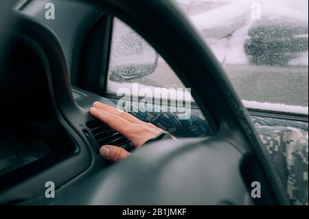 la main de l'homme à peau claire chauffe près d'un chauffage de voiture dans l'intérieur noir de la voiture contre le fond d'une fenêtre dans la neige. Préchauffage du Banque D'Images